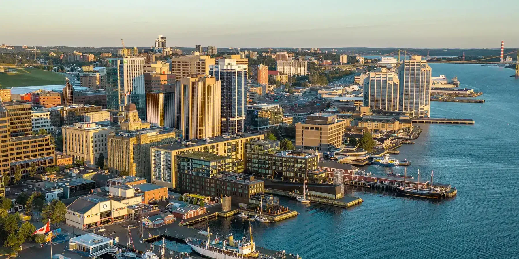 Aerial view of downtown Halifax at golden hour, looking along the waterfront boardwalk and harbour toward the MacKay Bridge.